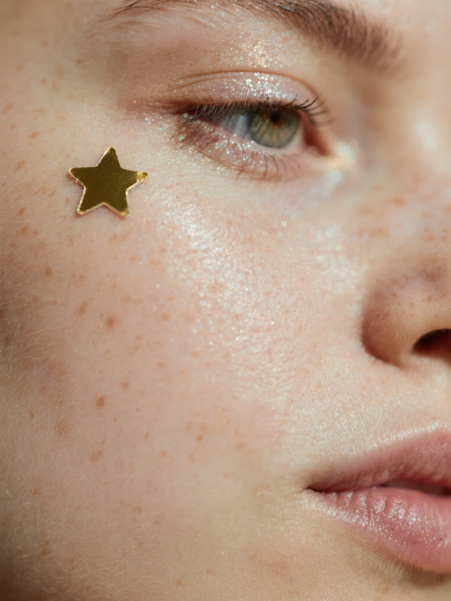 A close-up of a freckled face with a gold star sticker and glitter makeup